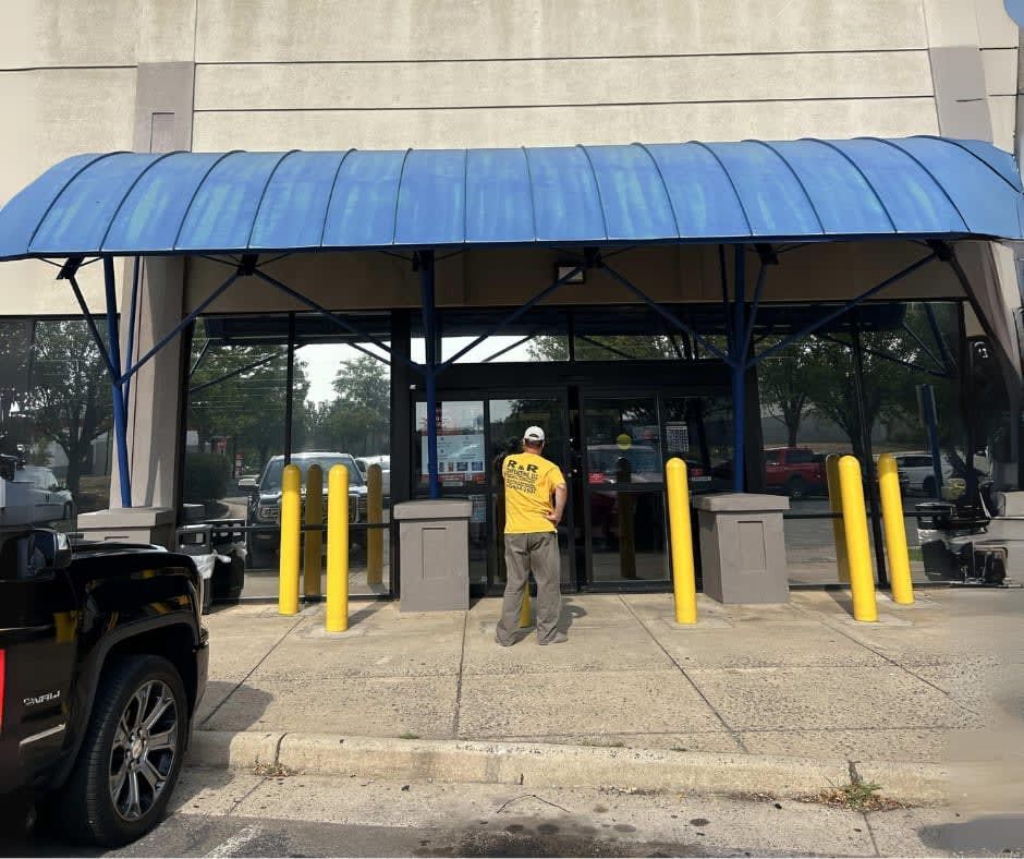 Person facing entrance of a building with blue awning, yellow poles, and reflective windows; a black vehicle is parked.