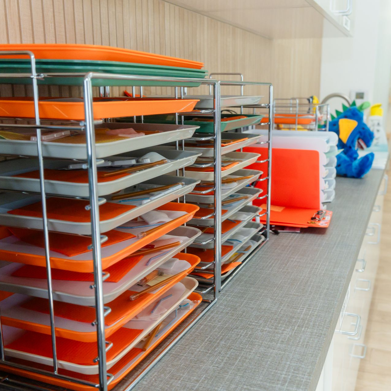 Stacked orange and gray trays on metal racks in a bright classroom, with colorful toys in the background