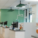 Modern dental clinic reception with white counters, mint green wall, and treatment chairs behind glass partitions