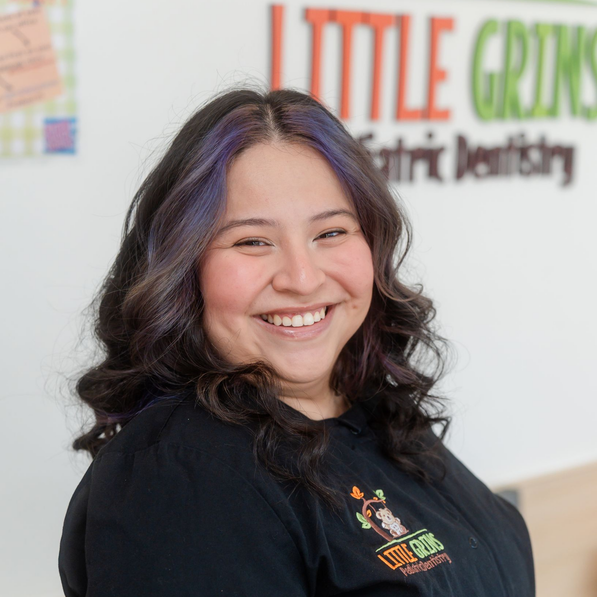 Smiling woman in a black shirt standing in front of a Little Grins Pediatric Dentistry sign