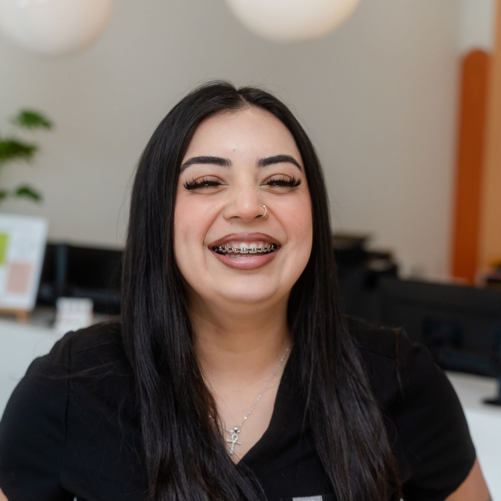 Smiling woman with long dark hair in a black top, seated indoors against a blurred office background