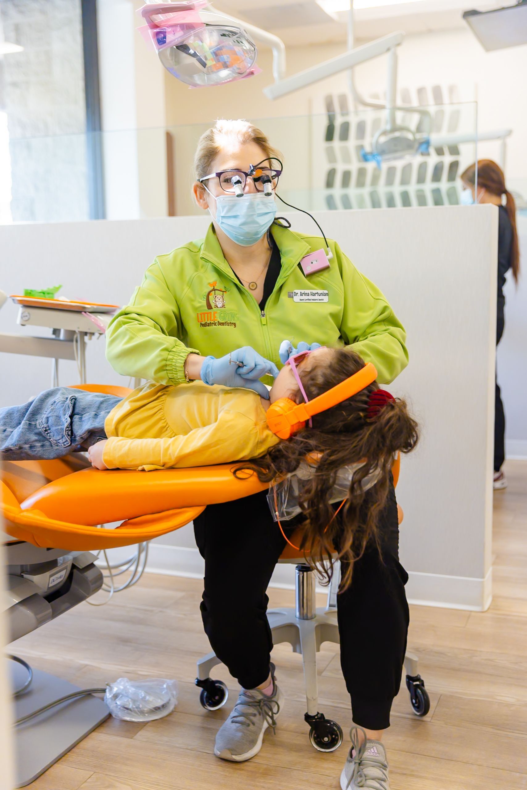 Medical worker in green jacket tending to a patient on an orange chair in a bright clinic.
