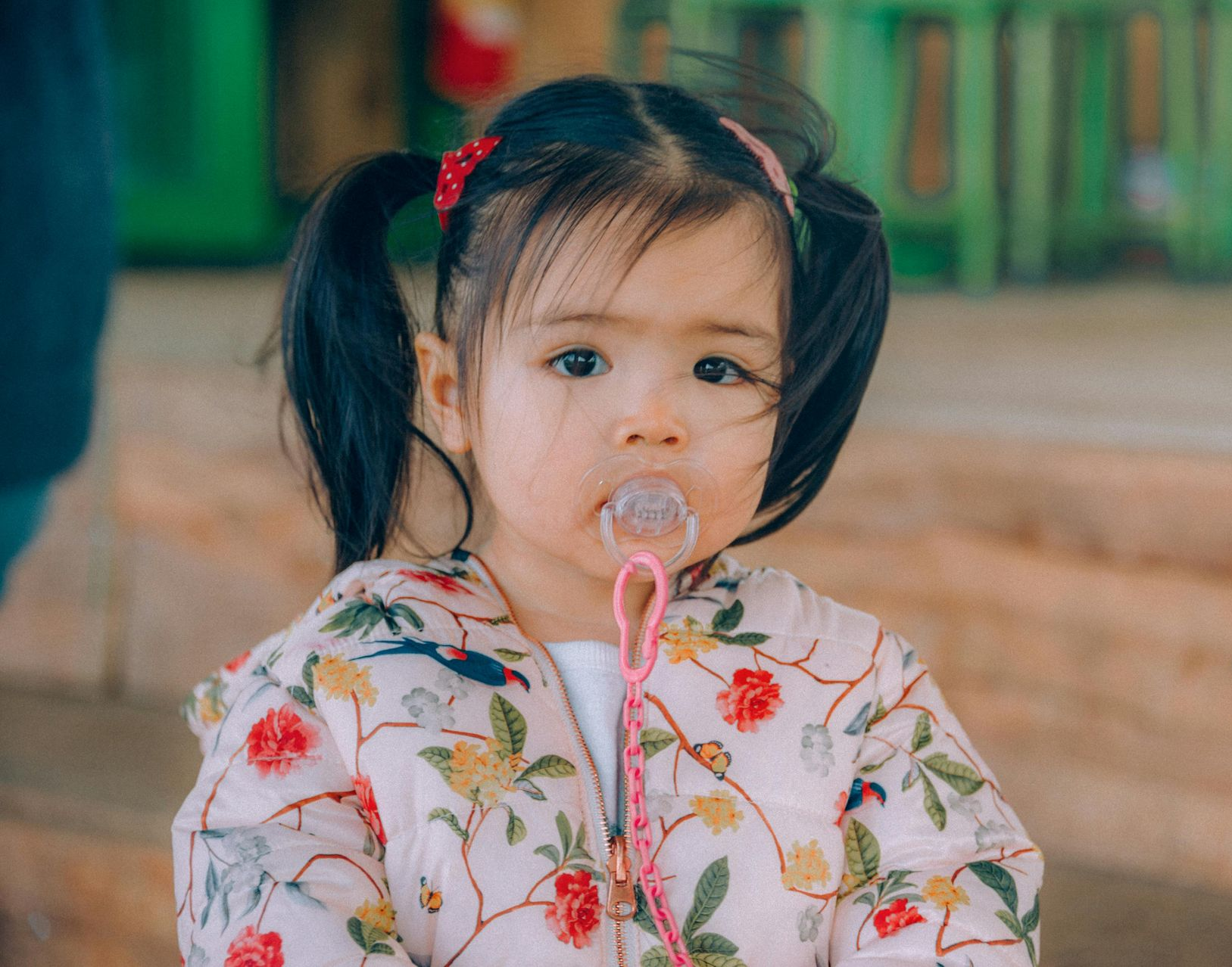 Child with pigtails sucking on a pink lollipop, wearing a floral shirt outdoors.