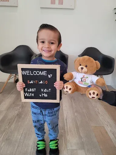 Child smiling in a room, holding a chalkboard sign that says “Welcome” beside a teddy bear.