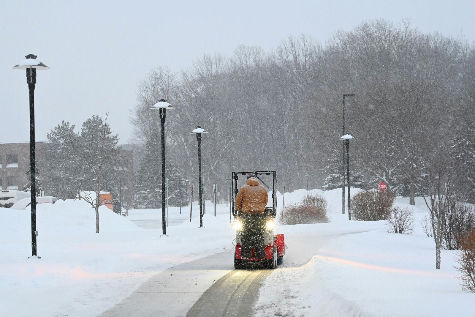 Snowplow clearing a curved path through deep snow in a park with lamp posts and trees.