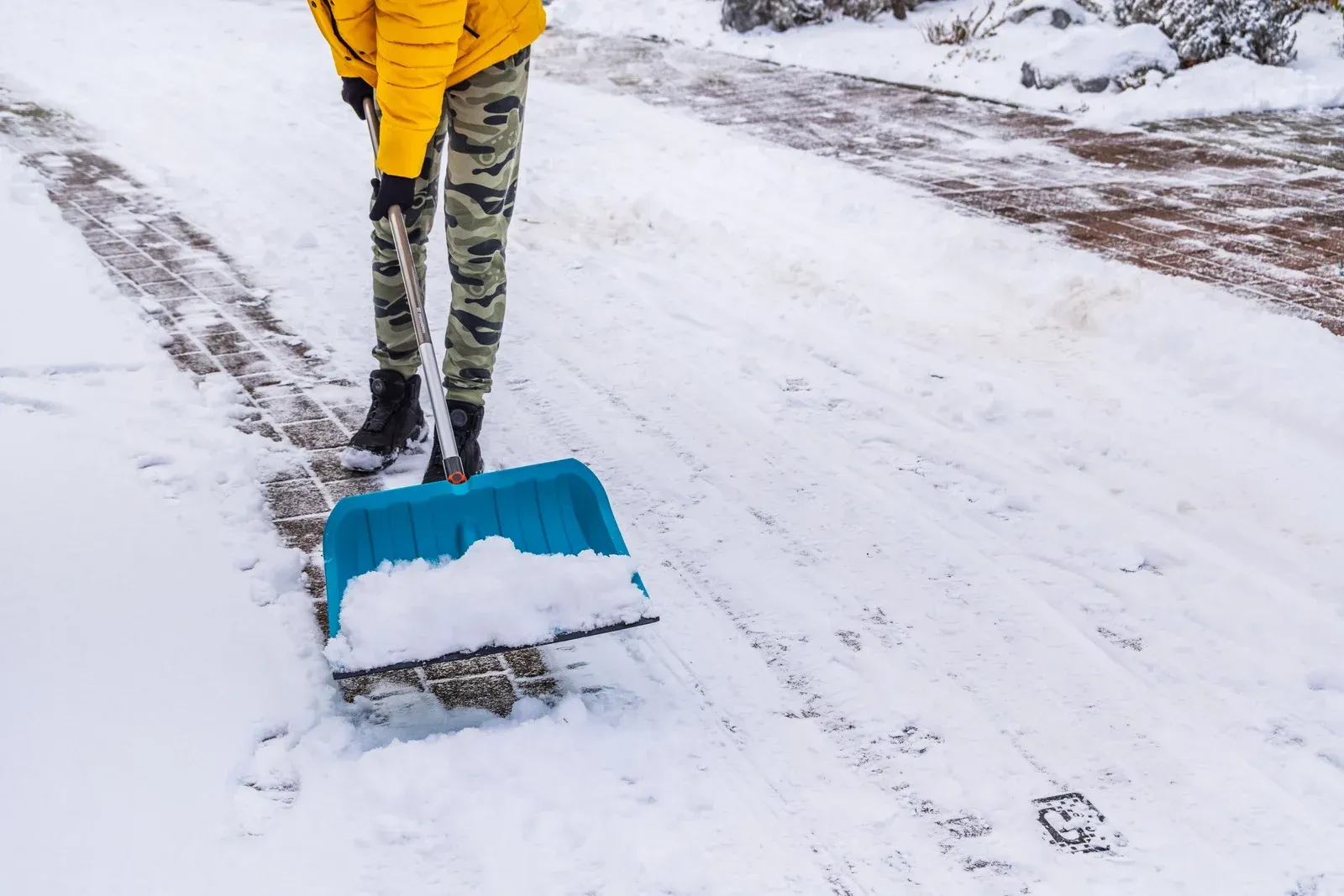 Person in yellow jacket shoveling snow with a blue snow shovel on a snowy road