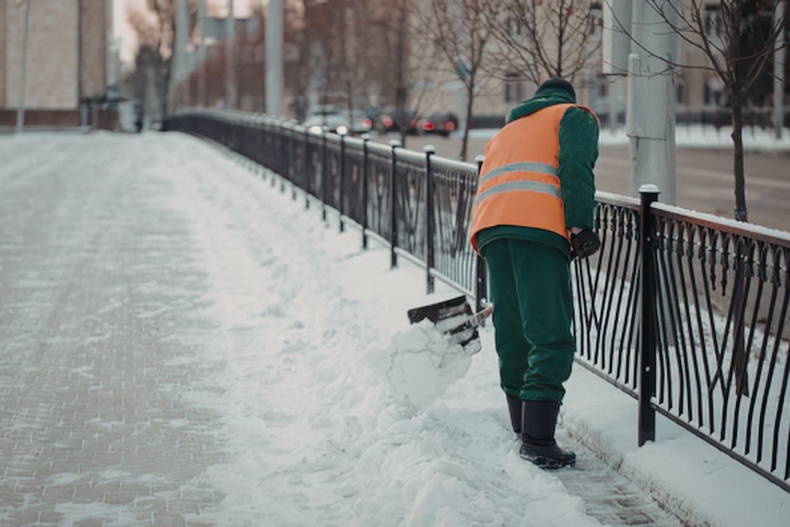 Worker in orange vest shoveling snow beside a black fence on a snowy sidewalk
