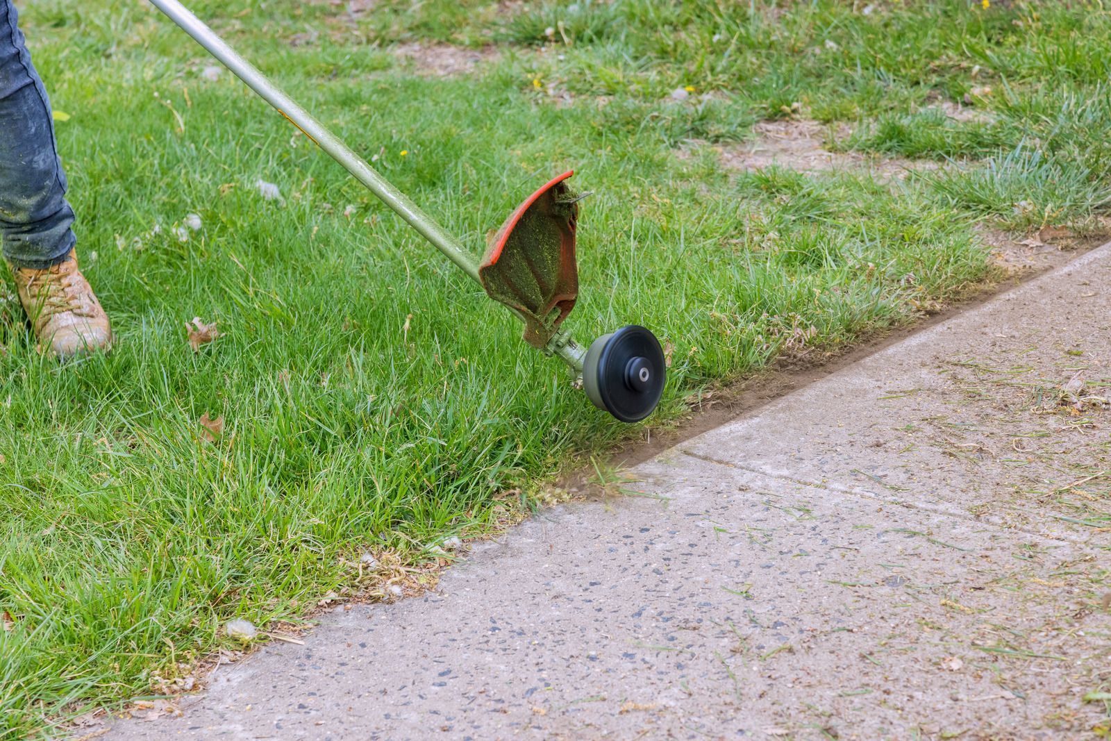 A weed trimmer cuts grass along a sidewalk edge, with a person’s leg visible on the left.