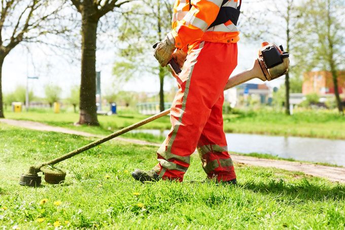 Worker using a string trimmer on grass beside a path, wearing orange safety pants and vest.