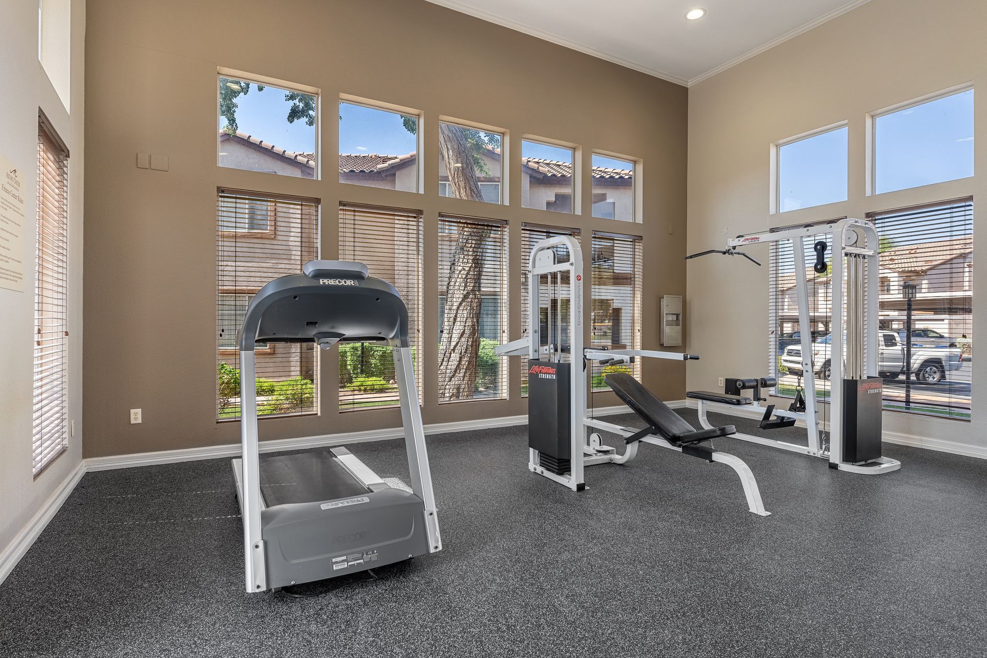 Bright gym room with treadmills, weight machines, and large windows overlooking trees and buildings