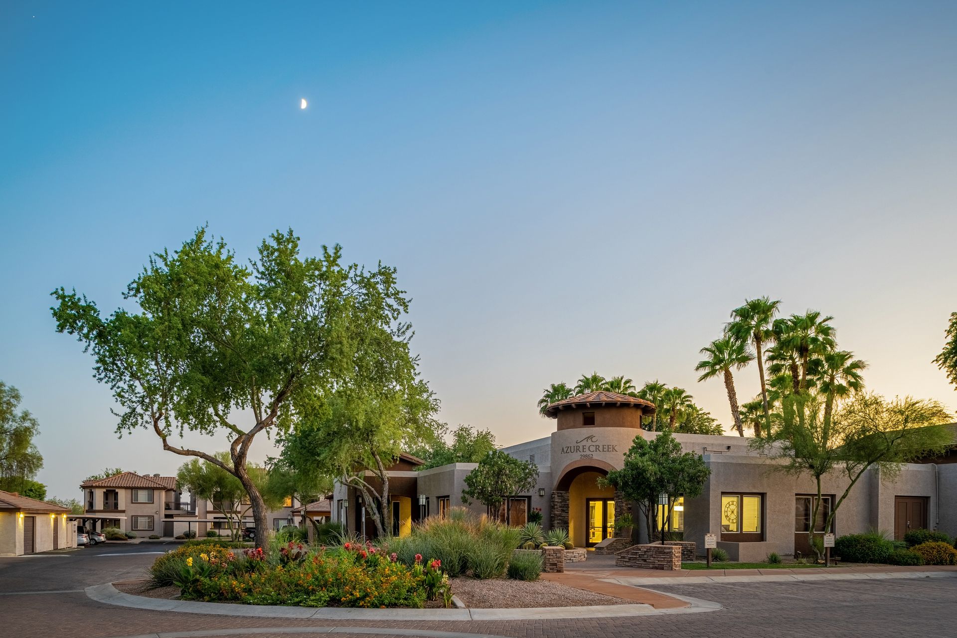 Landscaped courtyard with a stucco building, lit windows, trees, and flower beds at dusk