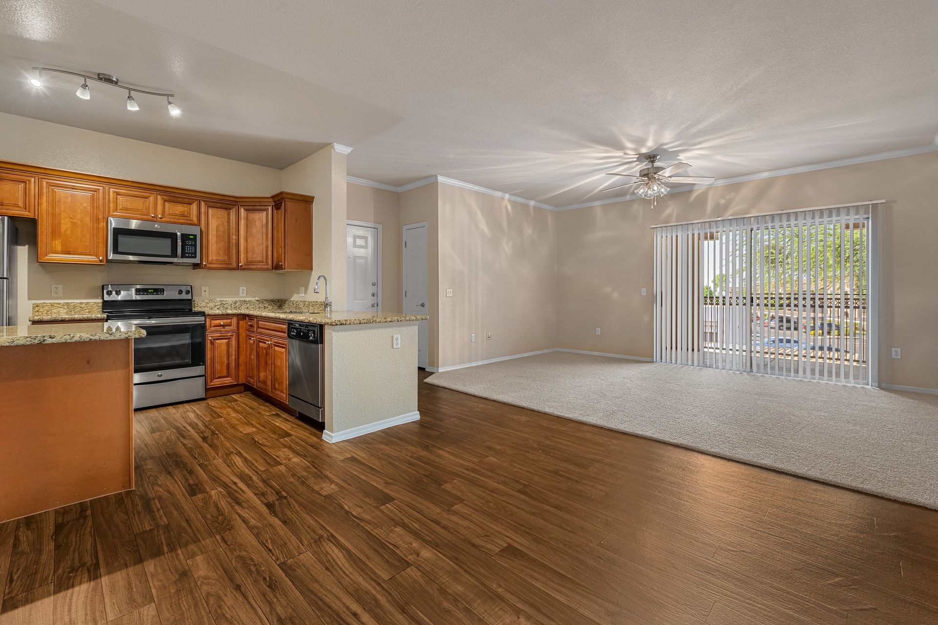Empty living room with kitchen, wood floors, beige carpet, and sliding glass doors to the outside