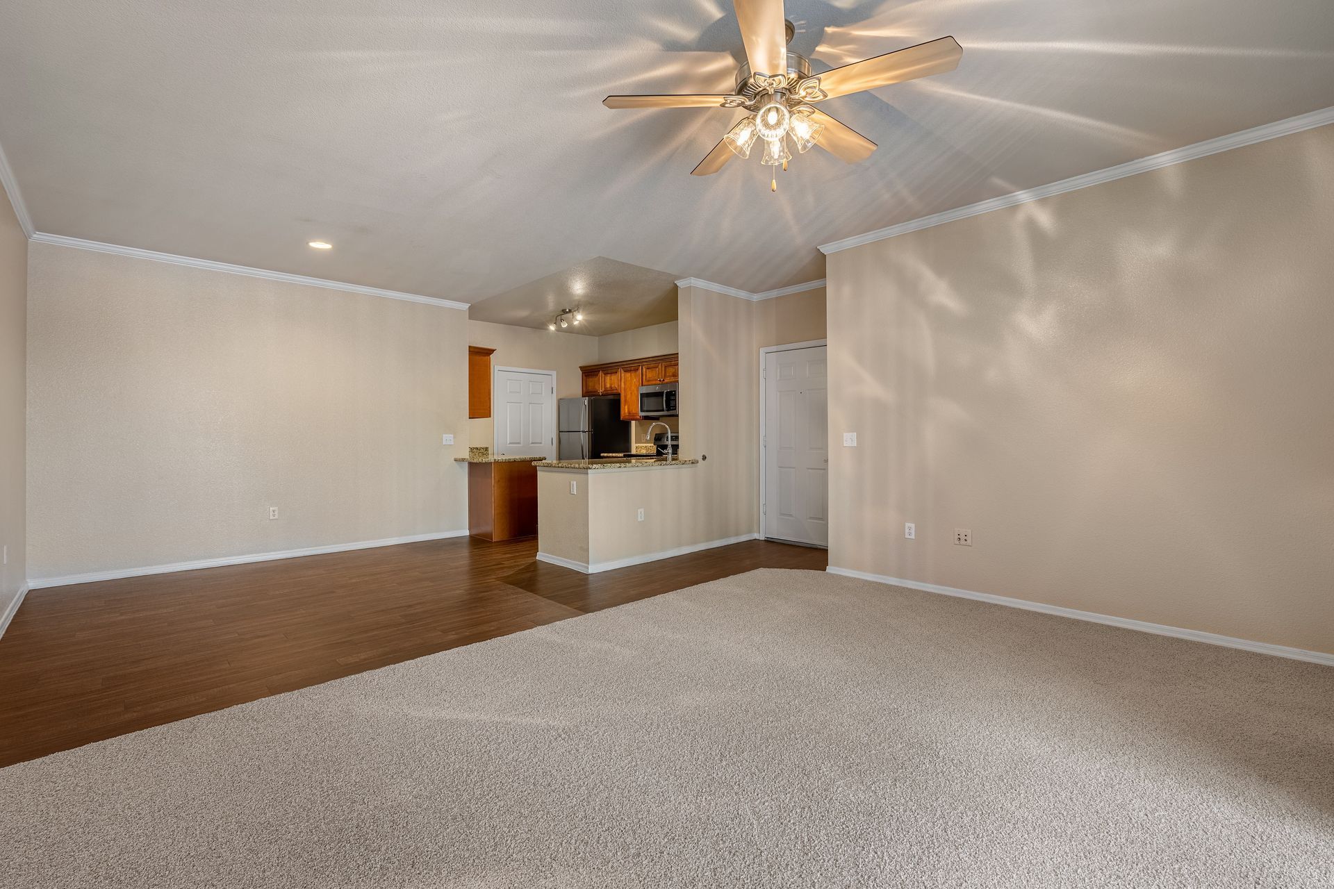 Empty carpeted living room with beige walls, ceiling fan, and open view into kitchen area