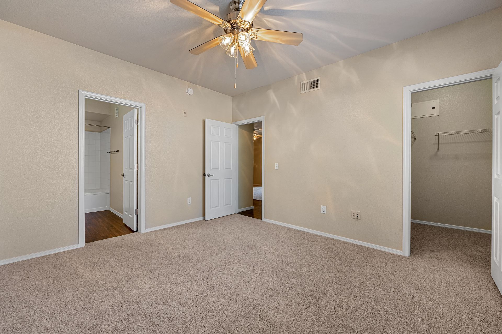 Empty carpeted bedroom with beige walls, ceiling fan, and three open doorways