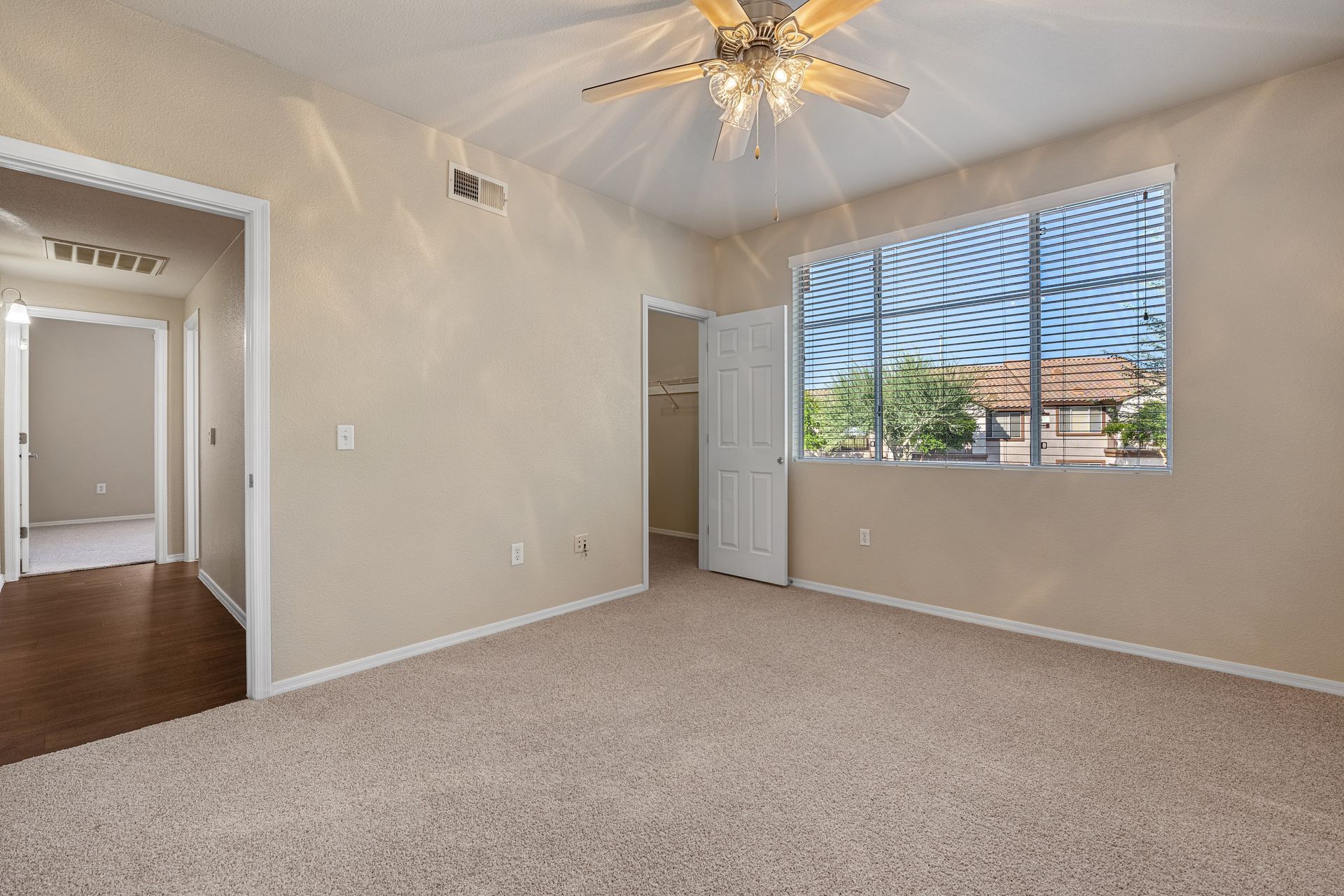 Empty beige bedroom with carpet, ceiling fan, and large window with white blinds.