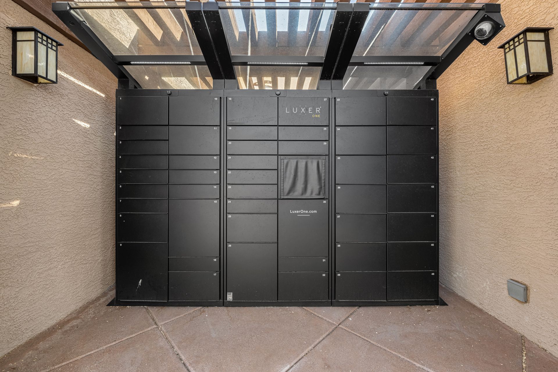 Modern black front gate in a narrow stucco entryway with wall lanterns and a glass awning.