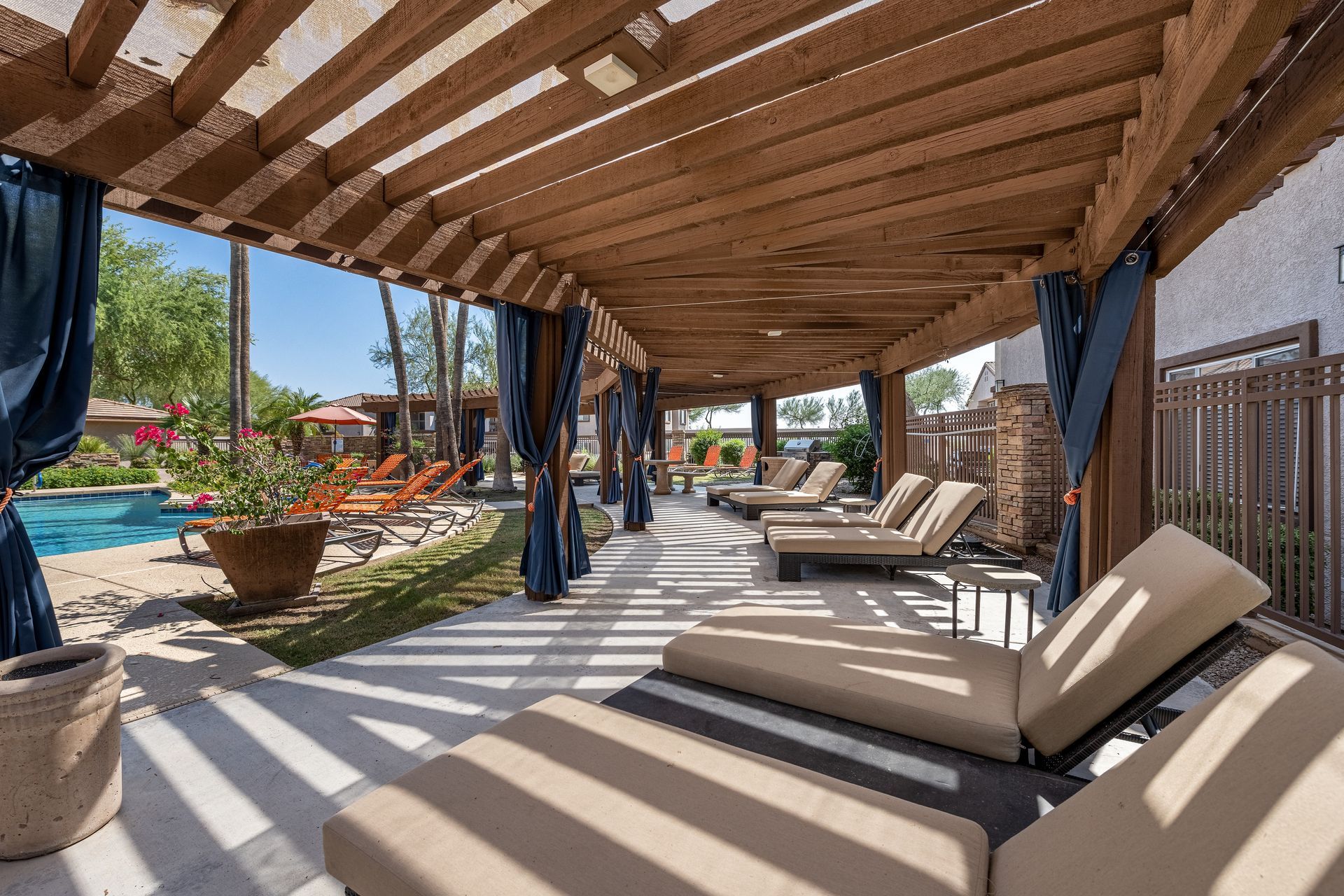 Covered outdoor patio with lounge chairs, striped shadows, potted plants, and a view of a pool area