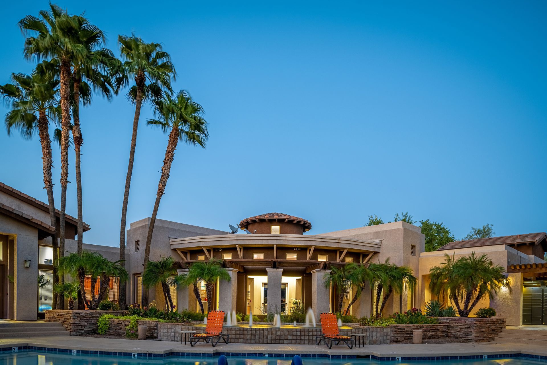 Modern resort courtyard with palm trees, a pool, and a lit entrance under a blue sky