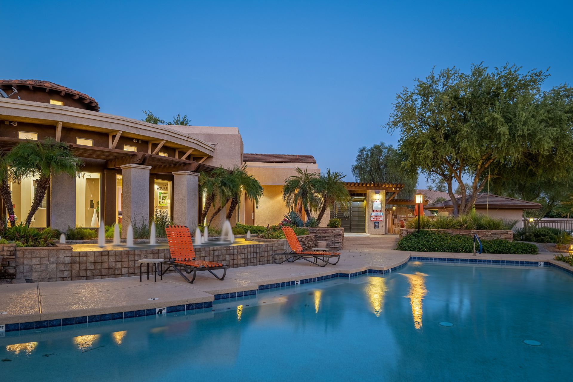 Luxury villa pool at dusk with lounge chairs, warm lights, and trees reflected in calm water
