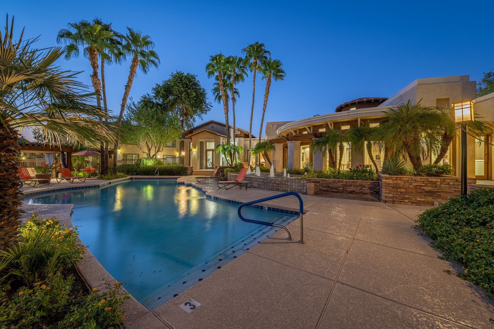 Resort-style outdoor pool at dusk with lit cabanas, palm trees, and a stone patio.