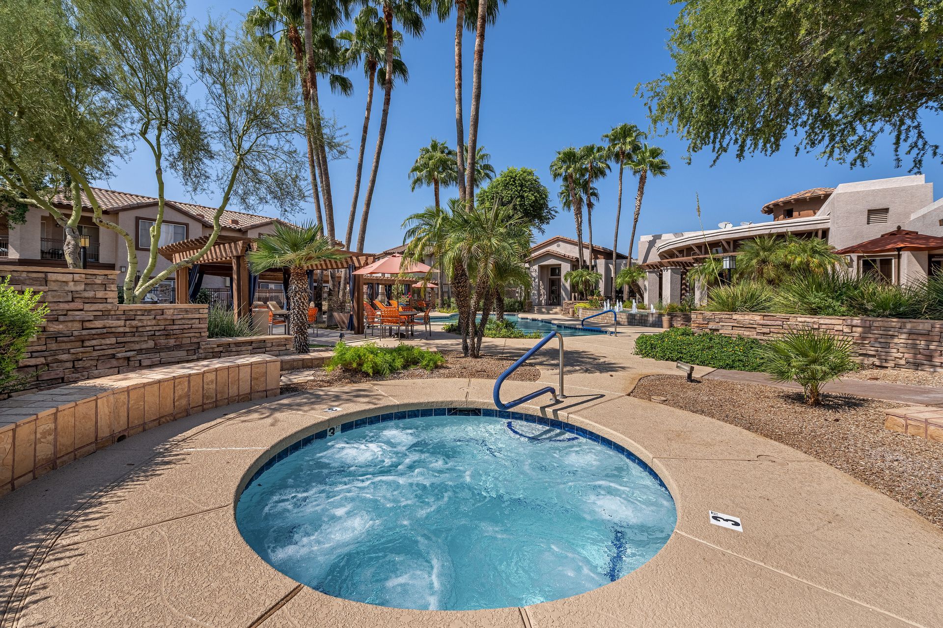 Resort courtyard with a small blue hot tub, palm trees, and tan stucco buildings under a bright sky