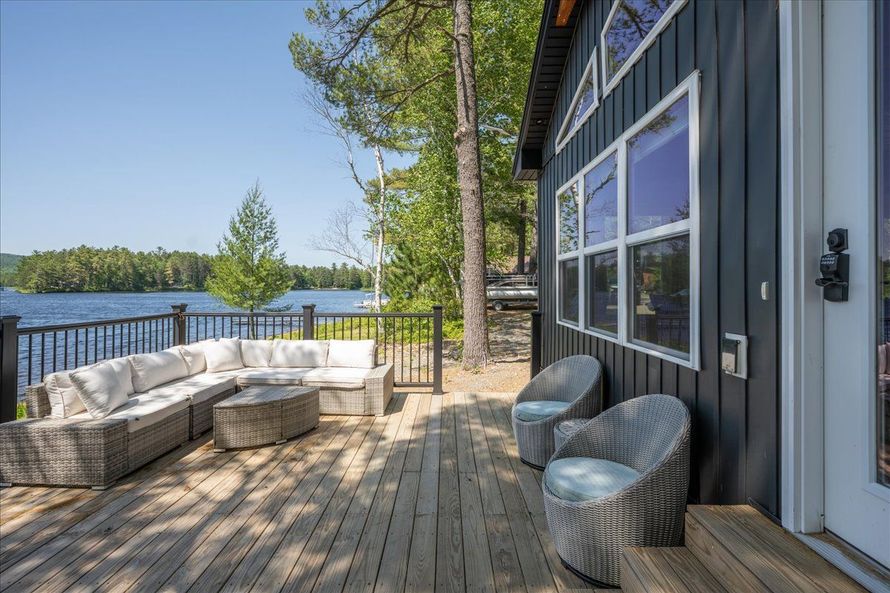 Lakefront deck with white sectional seating, wicker chairs, and a dark gray house under a blue sky