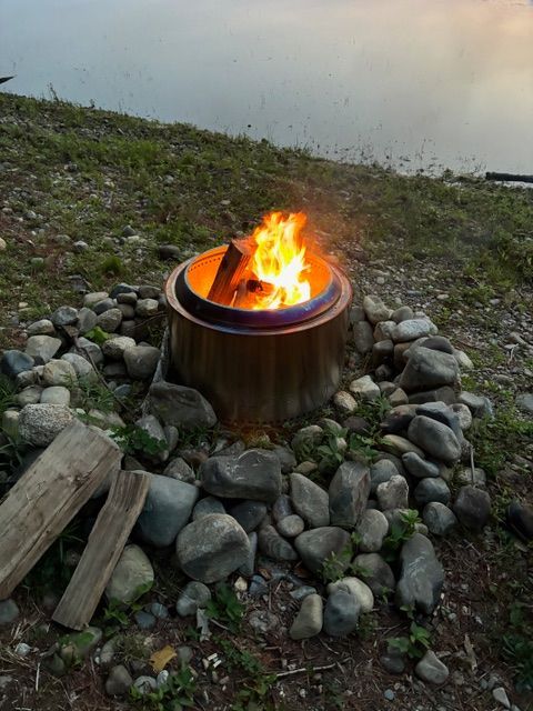 Fire burning in a metal fire pit surrounded by stones on a grassy, rocky hillside.