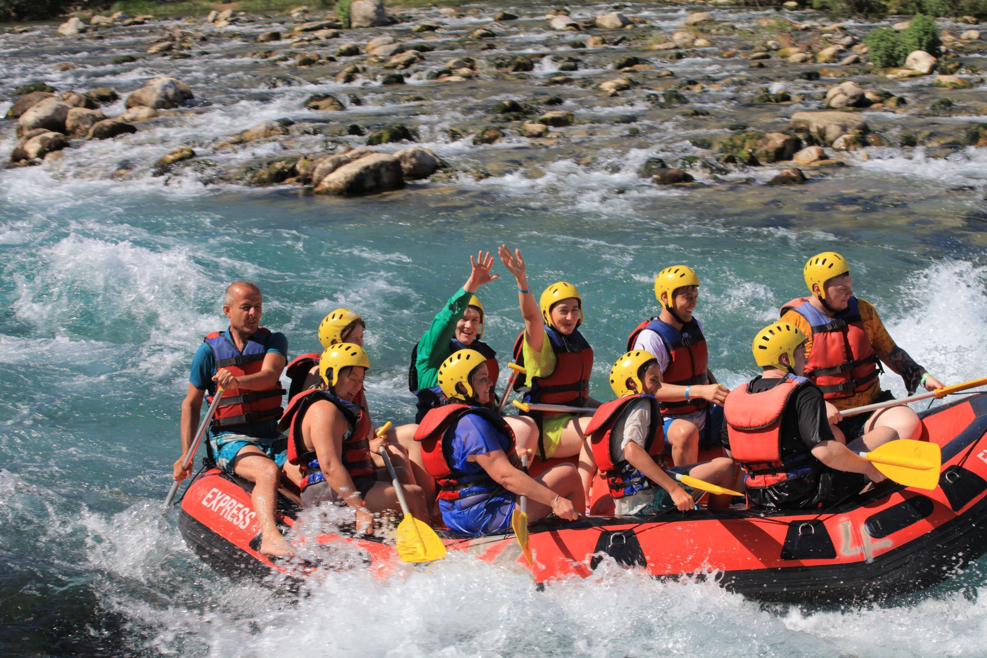 Group whitewater rafting in a red inflatable raft, with paddlers in yellow helmets splashing through rapids