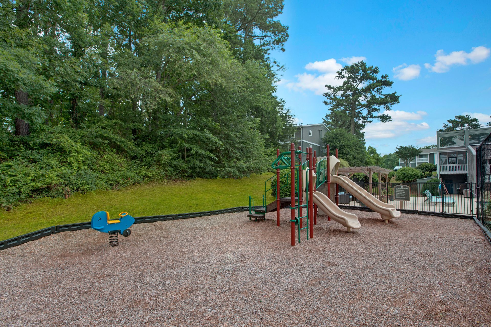 Children’s playground with slides and climbing structures on a gravel surface beside trees and buildings