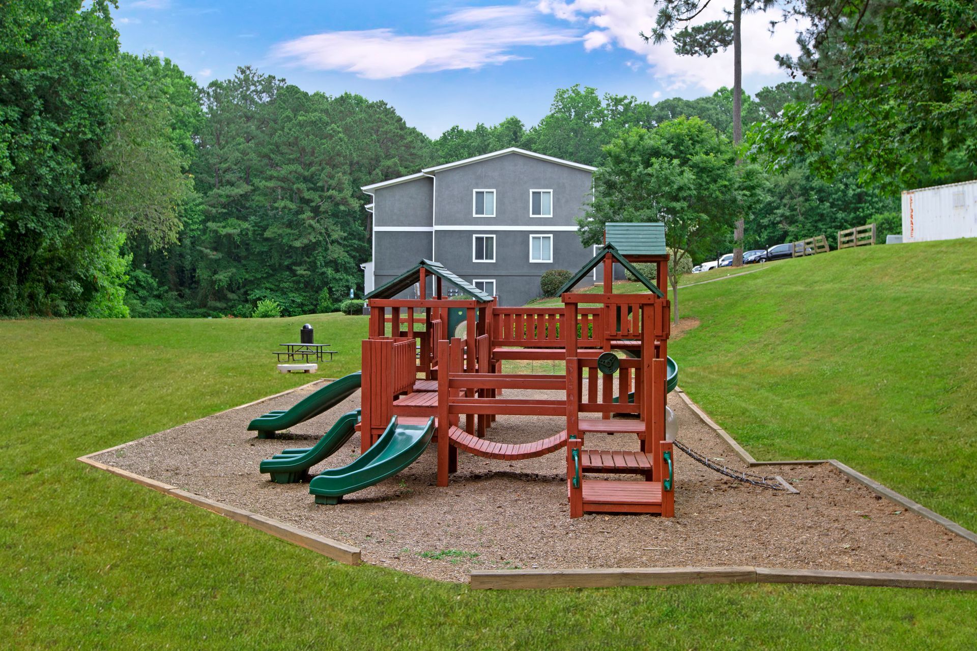 Playground with red and green slides on a grassy hill, with a gray house and trees in the background