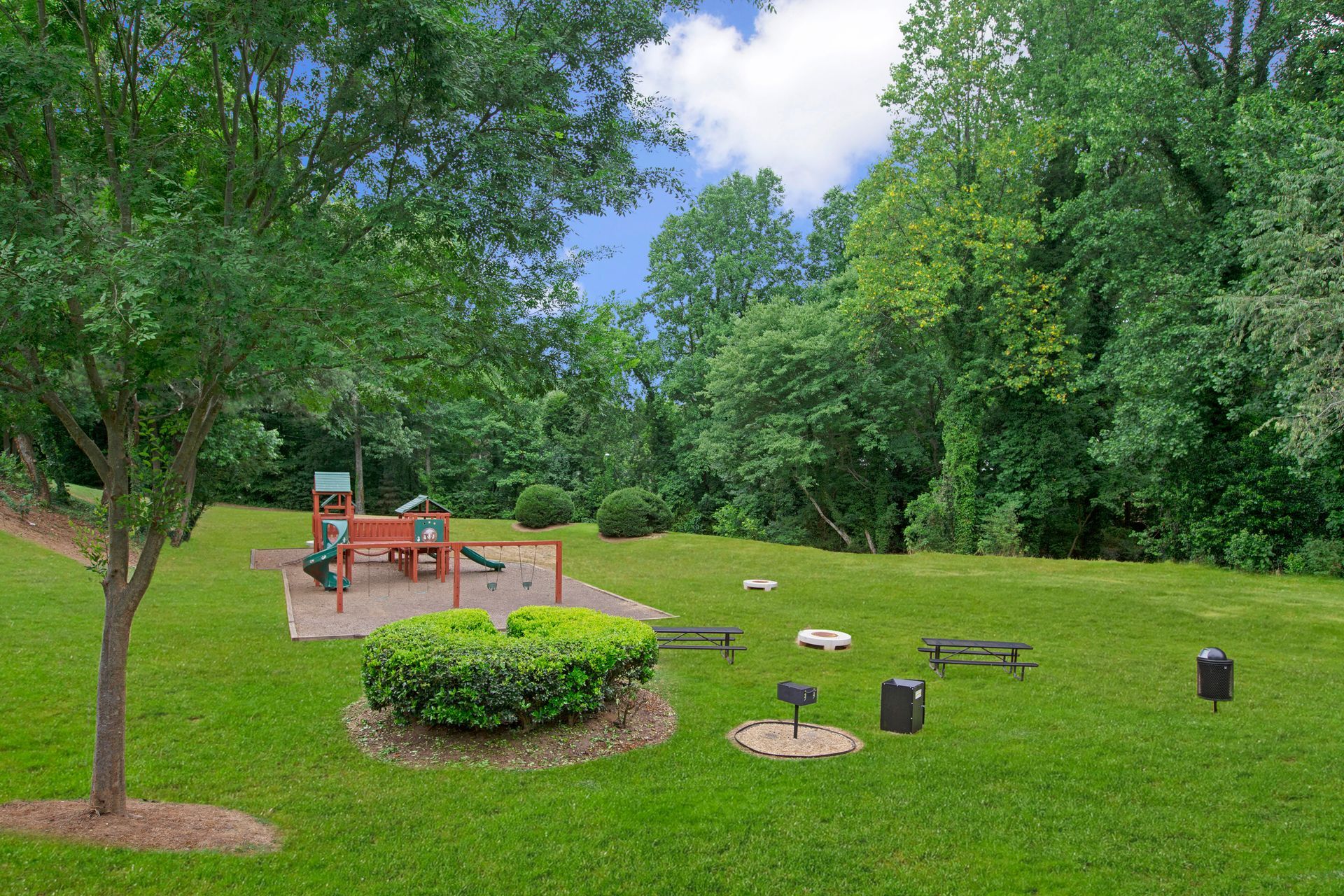 Wooded park with green lawn, picnic tables, fire pit, and a small shelter under trees