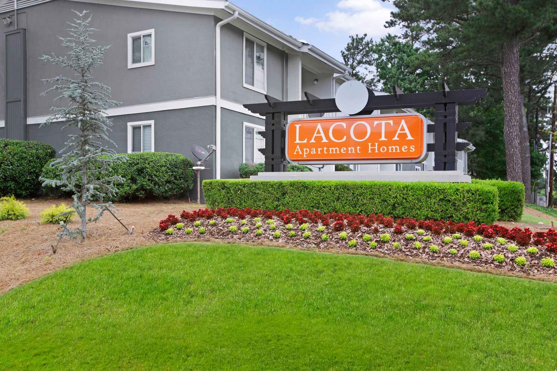Apartment complex sign in landscaped front yard: “Lacota Apartments” on orange panel beside gray building.