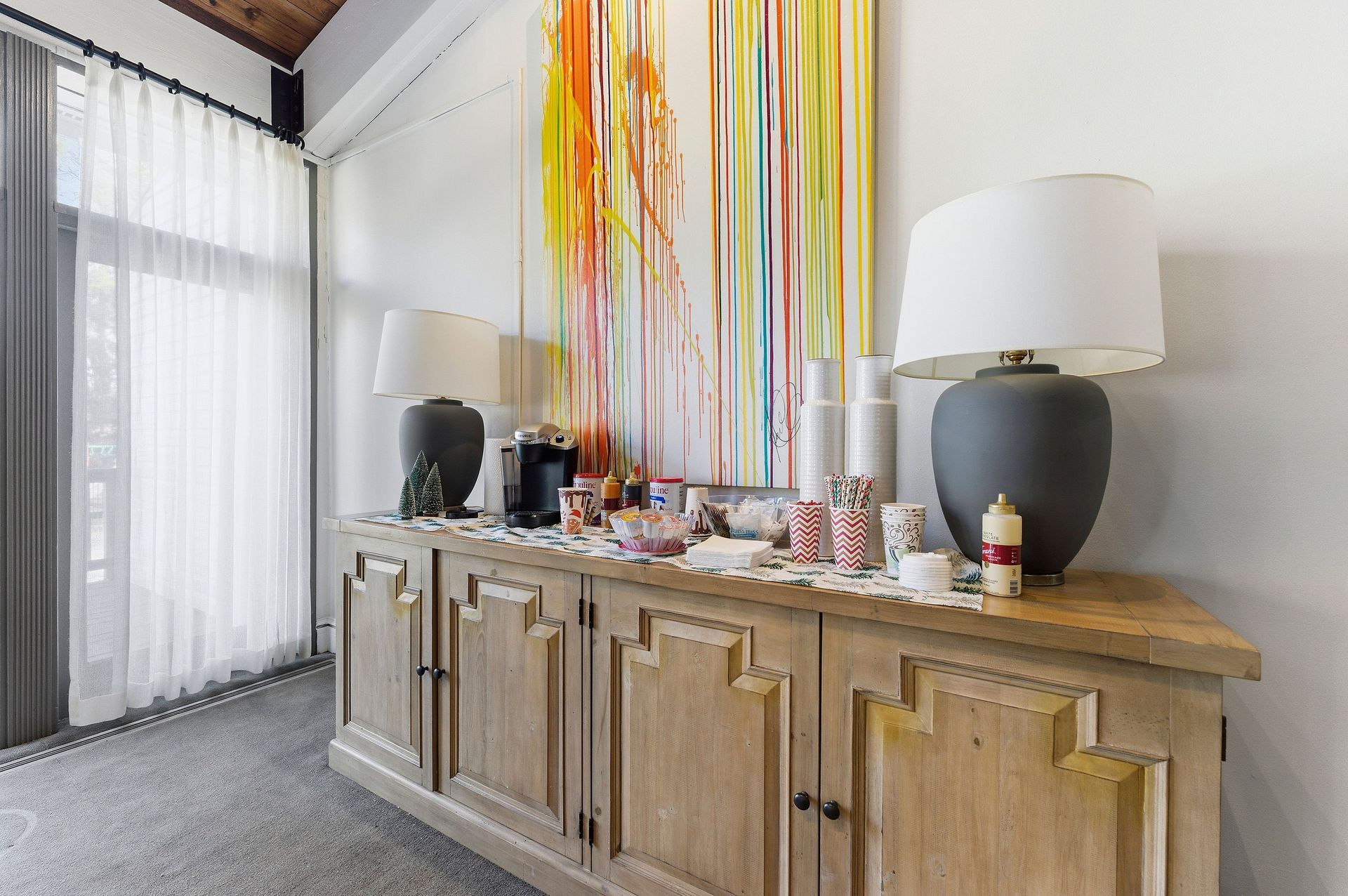 Living room sideboard with colorful ribbon art, lamps, and decor beneath tall windows