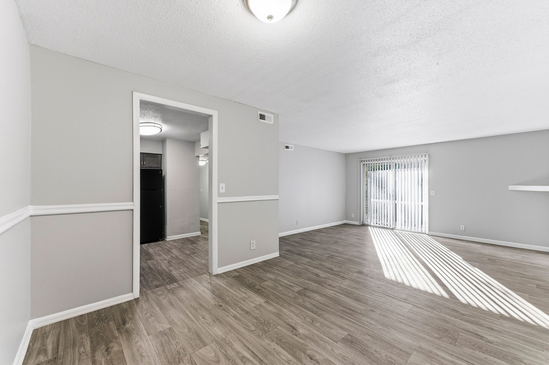 Empty living room with gray walls, wood flooring, and sunlight streaming through vertical blinds.