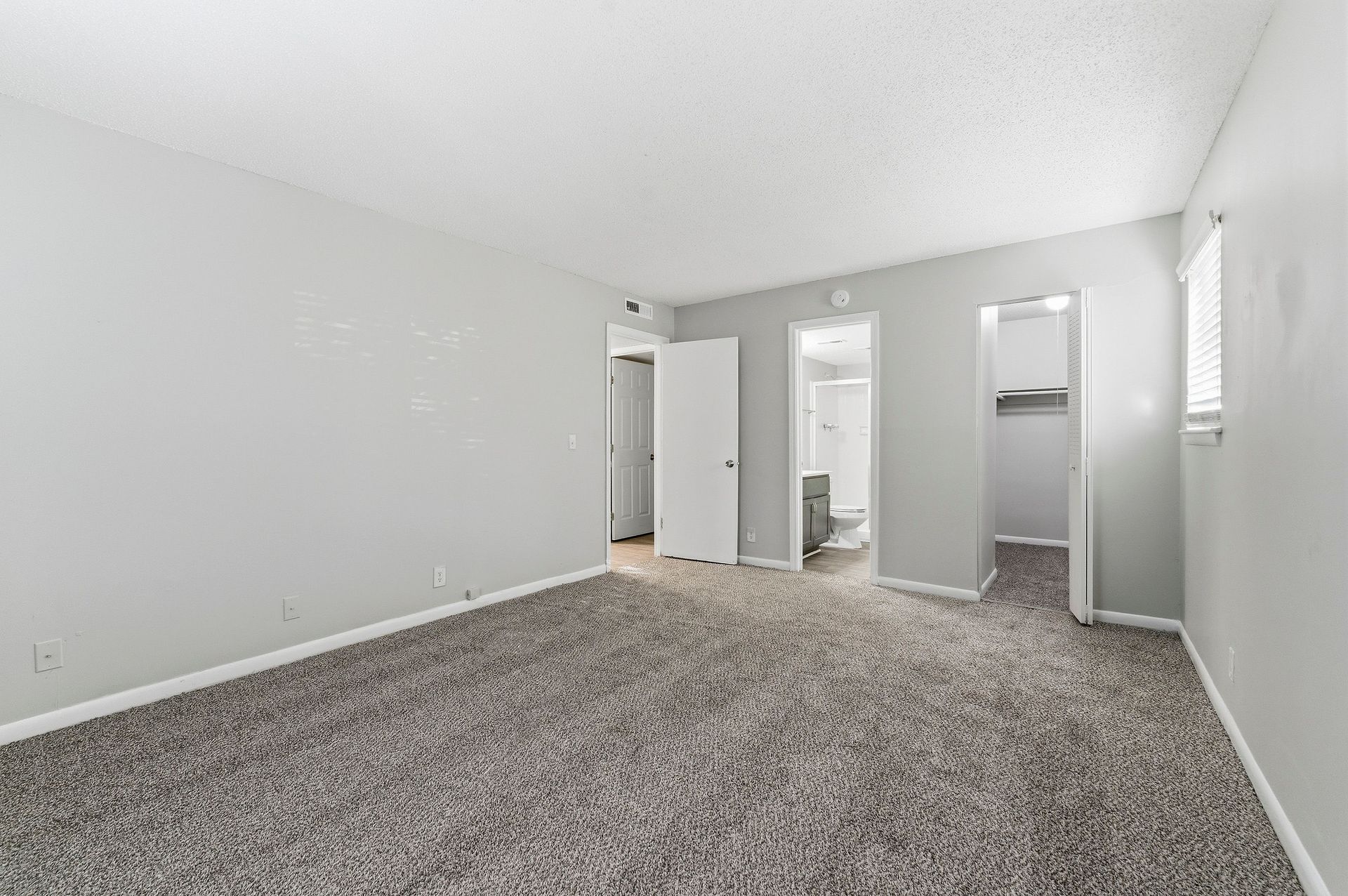 Empty carpeted bedroom with gray walls, white trim, and two open doorways leading to a bathroom and closet.