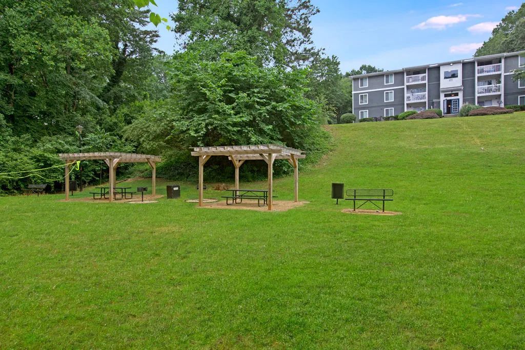 Grassy park with two wooden gazebos, a small picnic table, and apartment buildings in the background