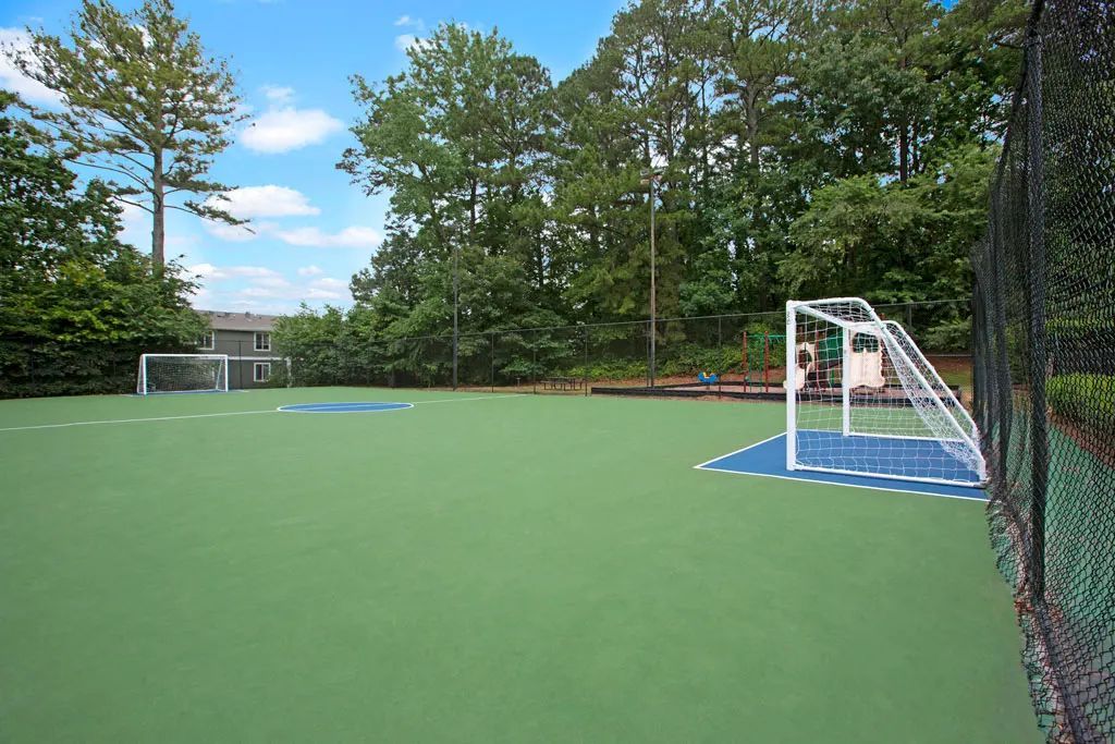 Empty outdoor sports court with a small soccer goal and green turf, bordered by trees and a netted fence