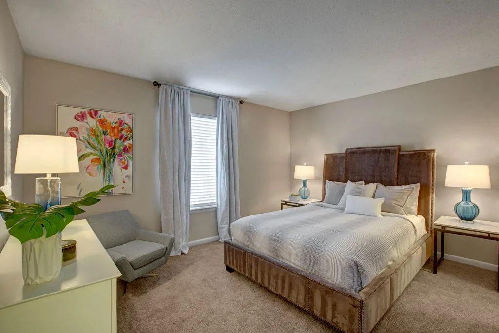 Bright bedroom with a wooden bed, two lamps, a gray chair, and floral wall art by a window