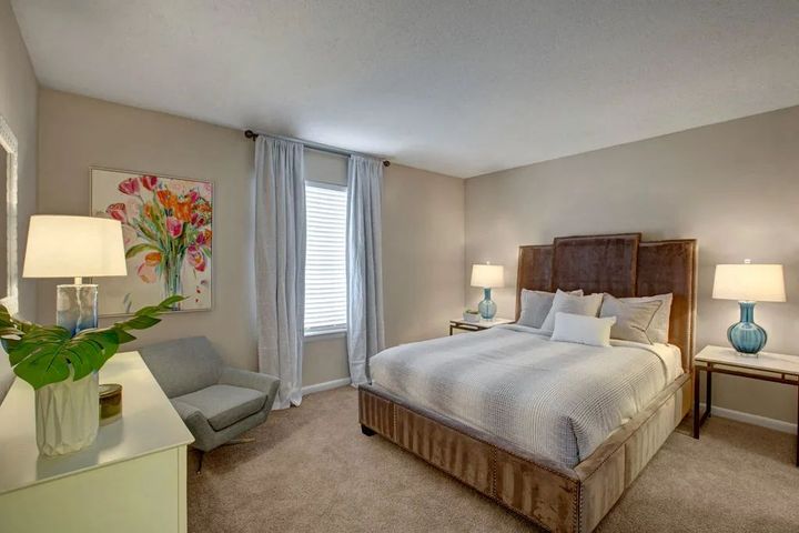 Bright bedroom with a wooden bed, two lamps, a gray chair, and floral wall art by a window