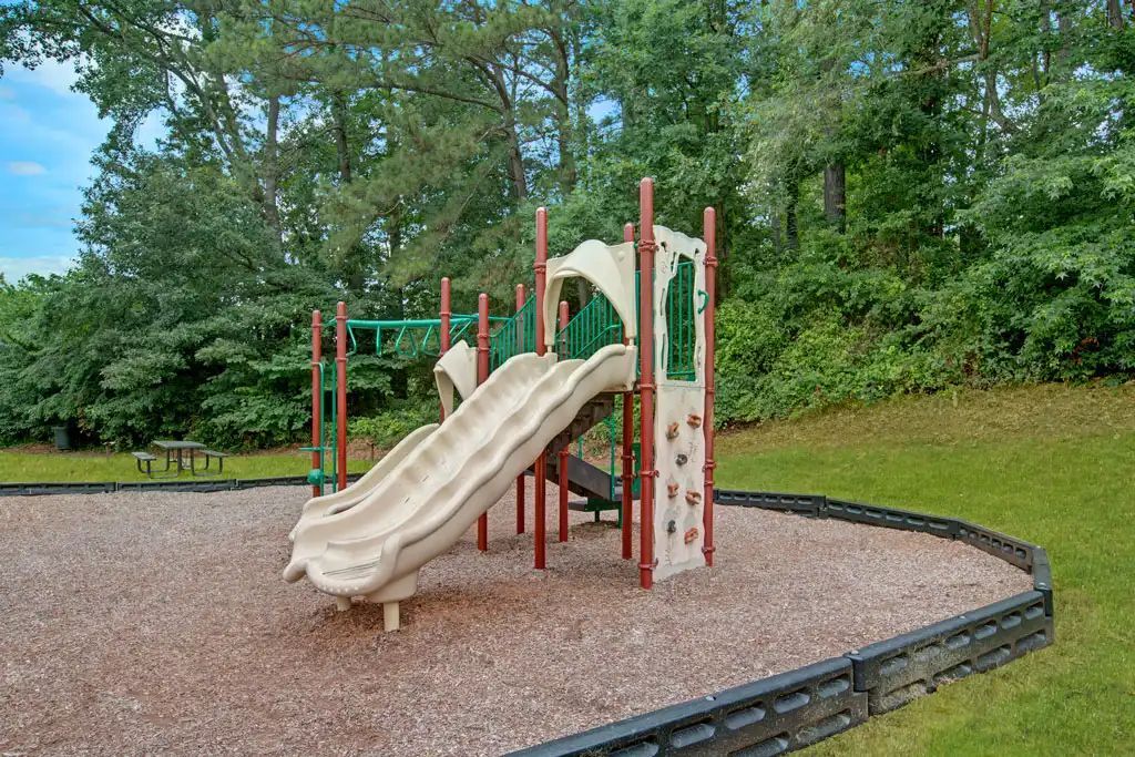 Empty playground with tan slide and red climbing structure on a mulch bed, bordered by grass and trees.