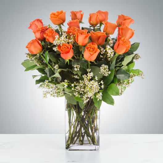 A square glass vase filled with bright orange roses and small white filler flowers against a plain gray background.