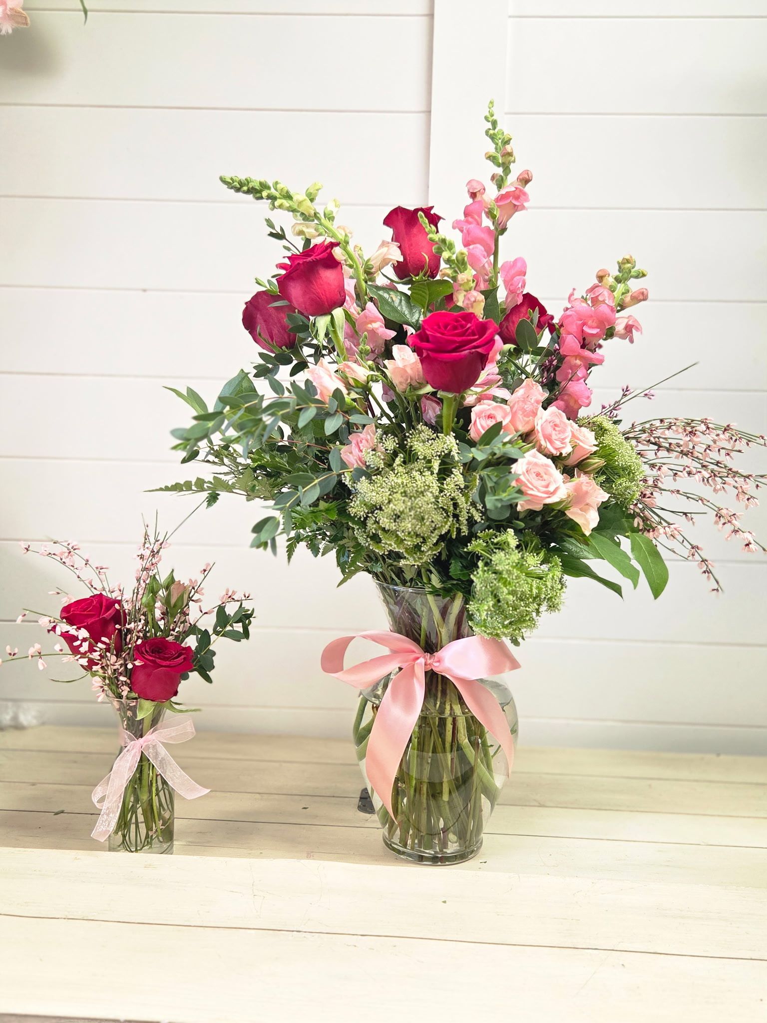 Two glass vases filled with deep pink roses, light pink snapdragons, and greenery, each tied with a light pink ribbon.