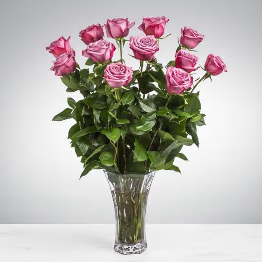 A bouquet of mauve roses in a clear glass vase against a plain grey background.