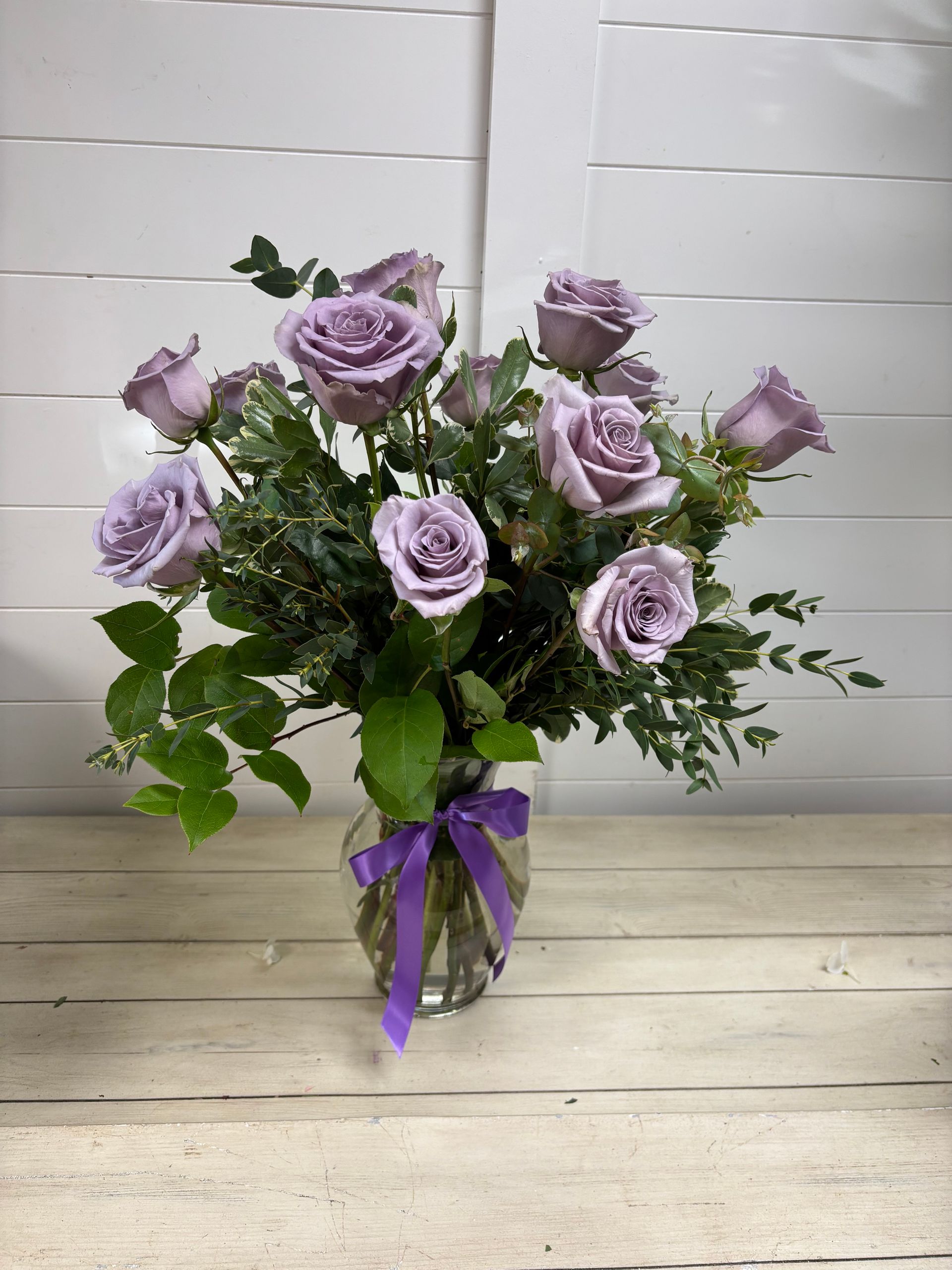 A vase of lavender roses with greenery, tied with a purple ribbon, sitting on a light wooden table against a white wall.