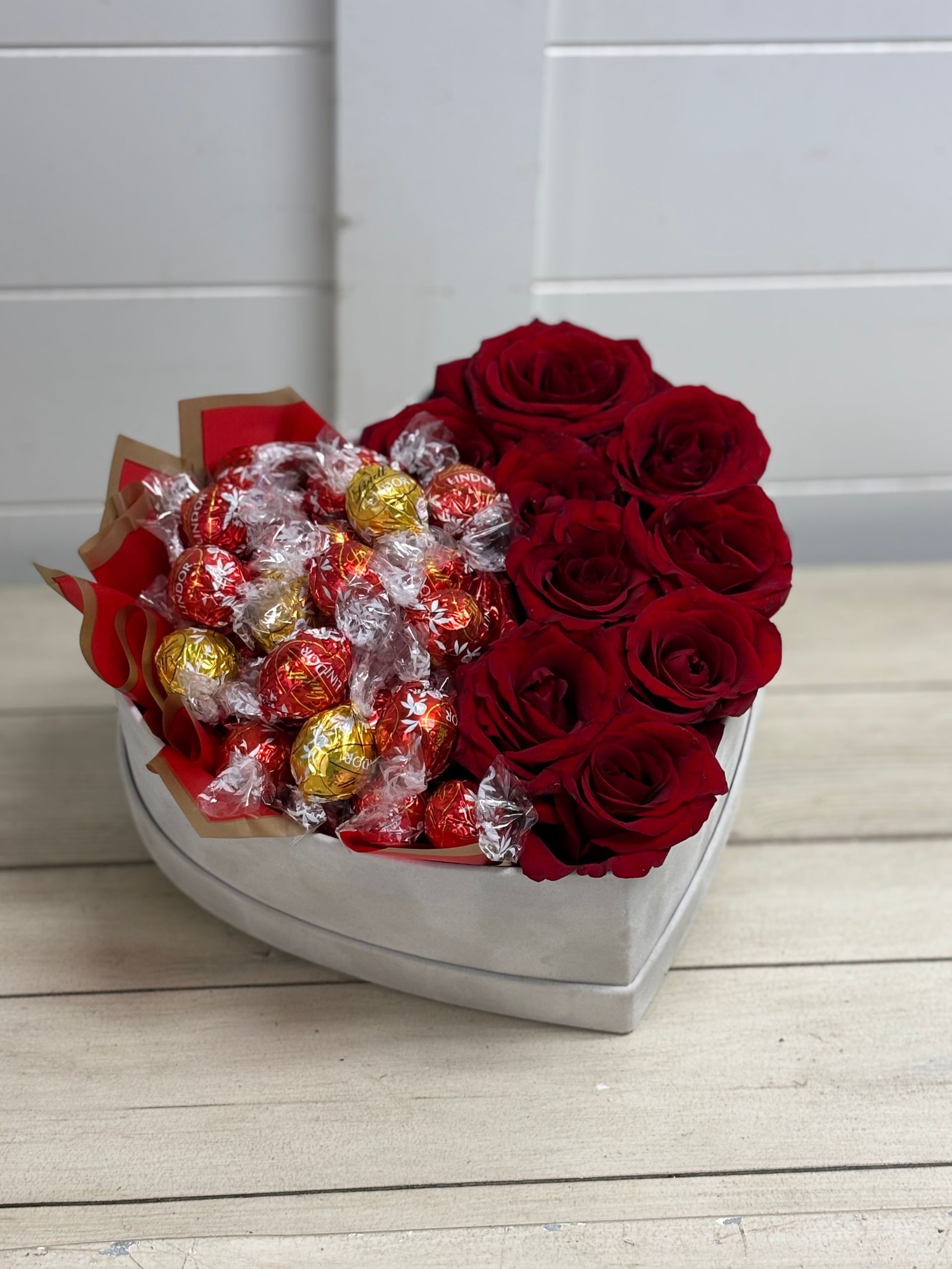 A heart-shaped box filled with red roses on one side and gold and red foil-wrapped chocolates on the other.
