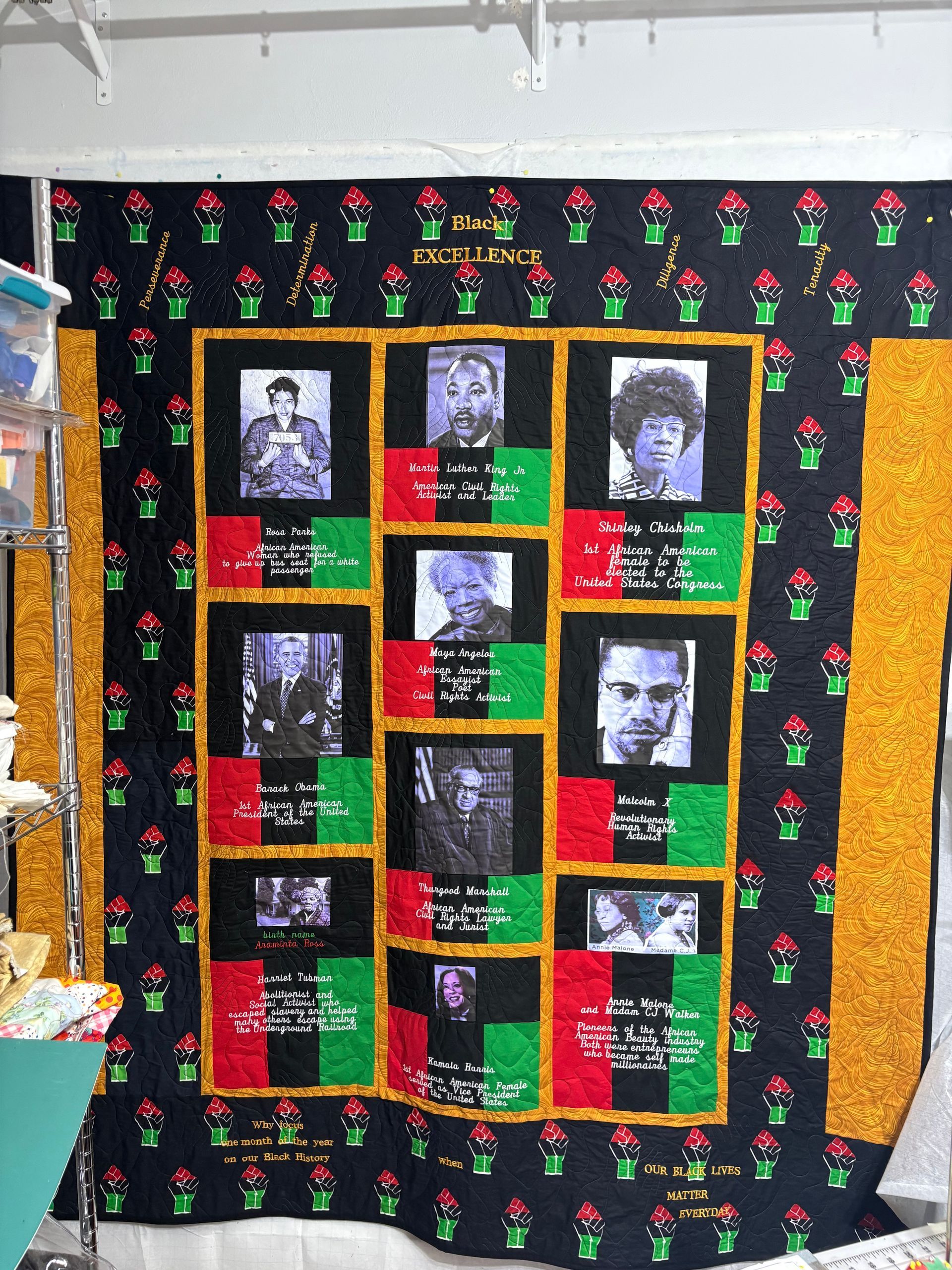 Black memorial-style quilt with nine portrait photos, red and green panels, and yellow trim on a wall
