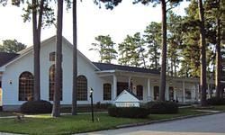 White building with arched windows and a porch, surrounded by tall trees and green grass.
