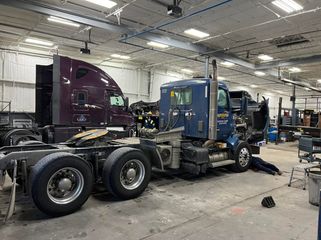 Blue semi-truck chassis in a repair shop, with a purple truck parked beside it.