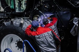 Worker inspecting a large tractor wheel and engine, wearing a red-and-gray jacket and blue cap