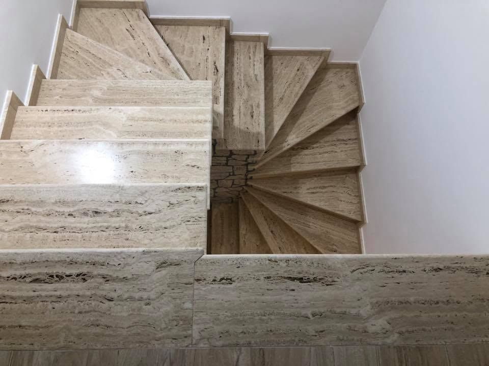 Scala a chiocciola con gradini in marmo beige e pareti bianche, vista dall'alto.